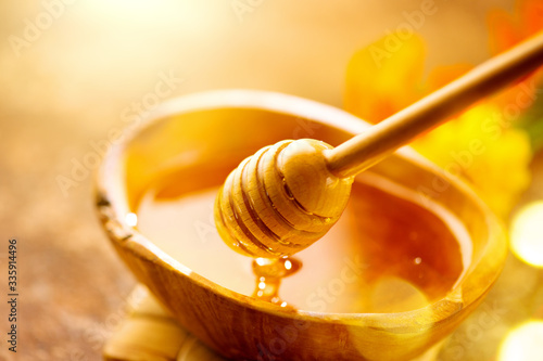 Honey dripping from honey dipper in wooden bowl. Close-up. Healthy organic Thick honey dipping from the wooden honey spoon, closeup. Flowers and jar on the table