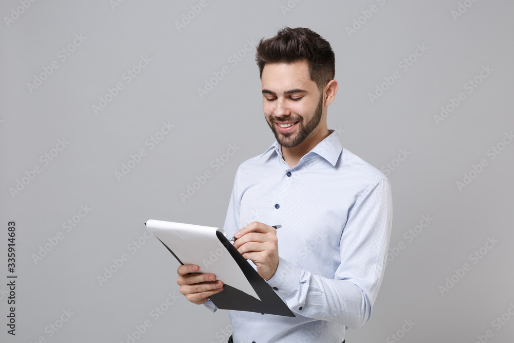 Smiling young unshaven business man in light shirt posing isolated on grey background. Achievement career wealth business concept. Mock up copy space. Hold clipboard with papers document write notes.