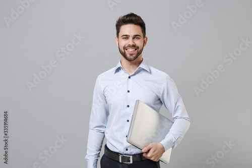 Smiling young unshaven business man in light shirt posing isolated on grey wall background studio portrait. Achievement career wealth business concept. Mock up copy space. Hold laptop pc computer.