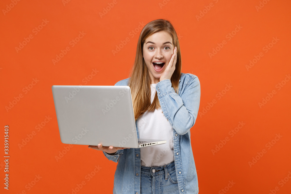 Surprised young woman girl in casual denim clothes posing isolated on orange wall background studio portrait. People lifestyle concept. Mock up copy space. Hold laptop pc computer, put hand on cheek. Surprised young woman girl in casual denim clothes posing isolated on orange wall background studio portrait. People lifestyle concept. Mock up copy space. Hold laptop pc computer, put hand on cheek.