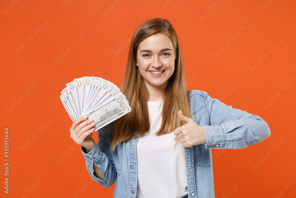 Smiling young woman girl in casual denim clothes posing isolated on orange wall background. People lifestyle concept. Mock up copy space. Hold fan of cash money in dollar banknotes, showing thumb up. Smiling young woman girl in casual denim clothes posing isolated on orange wall background. People lifestyle concept. Mock up copy space. Hold fan of cash money in dollar banknotes, showing thumb up.