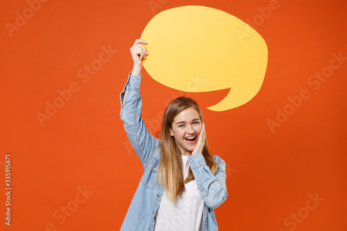 Funny young woman girl in casual denim clothes posing isolated on orange background. People lifestyle concept. Mock up copy space. Hold yellow empty blank Say cloud, speech bubble, put hand on cheek. © ViDi Studio