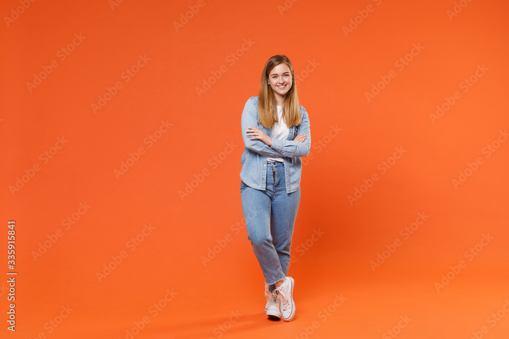 Smiling young woman girl in casual denim clothes posing isolated on bright orange wall background studio portrait. People sincere emotions lifestyle concept. Mock up copy space. Holding hands crossed. Smiling young woman girl in casual denim clothes posing isolated on bright orange wall background studio portrait. People sincere emotions lifestyle concept. Mock up copy space. Holding hands crossed.