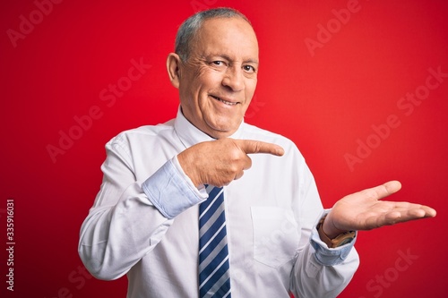 Senior handsome businessman wearing elegant tie standing over isolated red background amazed and smiling to the camera while presenting with hand and pointing with finger.