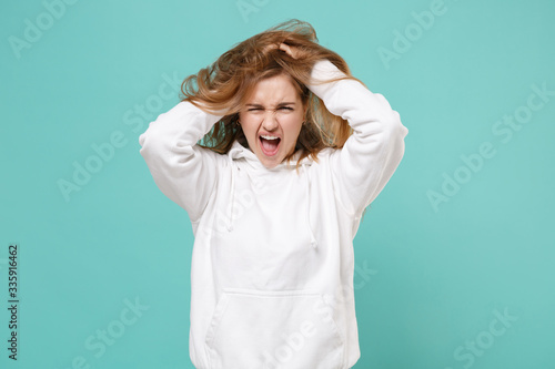 Crazy young woman girl in casual white hoodie posing isolated on blue turquoise wall background studio portrait. People emotions lifestyle concept. Mock up copy space. Put hands on head ruffling hair.