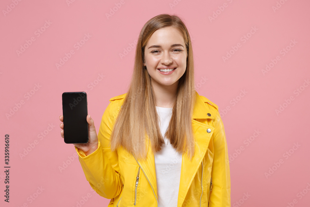 Smiling young woman girl in yellow leather jacket posing isolated on pastel pink background studio portrait. People lifestyle concept. Mock up copy space. Hold mobile phone with blank empty screen. Smiling young woman girl in yellow leather jacket posing isolated on pastel pink background studio portrait. People lifestyle concept. Mock up copy space. Hold mobile phone with blank empty screen.