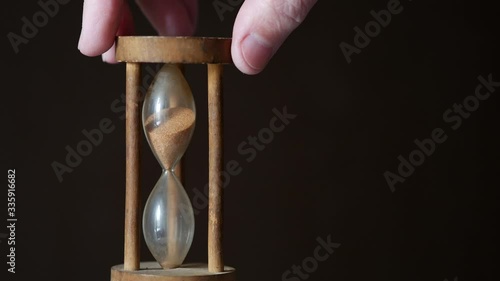 A male hand flips a vintage hourglass on a brown dark background. Unique wooden glass hourglass counts down the flowing sand. Countdown timer. Concept of passing time. B-roll for captions. Copy space