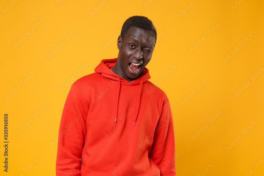 Cheerful young african american man guy in red streetwear hoodie posing isolated on yellow wall background studio portrait. People sincere emotions lifestyle concept. Mock up copy space. Blinking. Cheerful young african american man guy in red streetwear hoodie posing isolated on yellow wall background studio portrait. People sincere emotions lifestyle concept. Mock up copy space. Blinking.