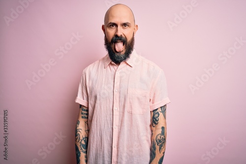 Handsome bald man with beard and tattoo wearing casual shirt over isolated pink background sticking tongue out happy with funny expression. Emotion concept.