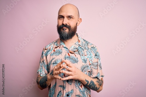 Handsome bald man with beard and tattoo wearing casual floral shirt over pink background Hands together and fingers crossed smiling relaxed and cheerful. Success and optimistic