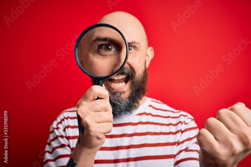 Handsome detective bald man with beard using magnifying glass over red background screaming proud and celebrating victory and success very excited, cheering emotion