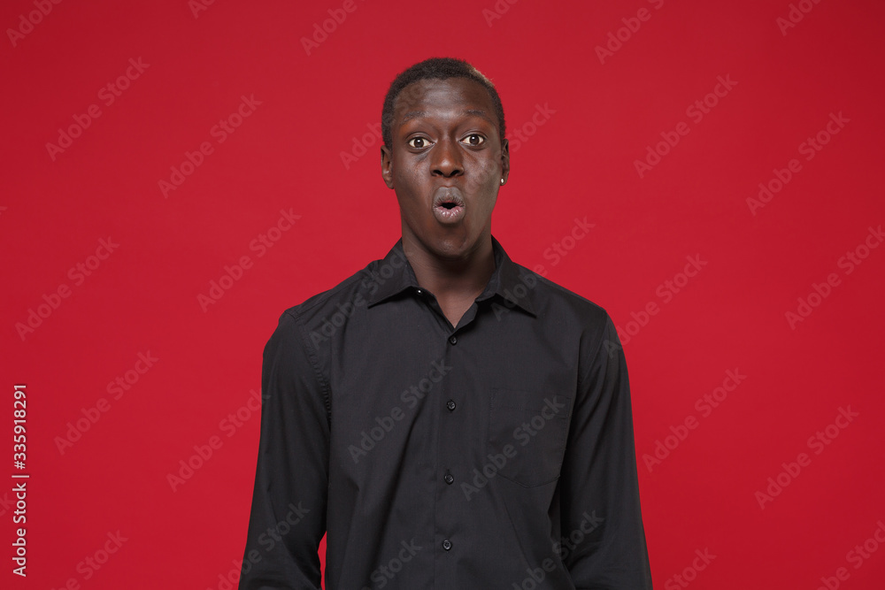 Shocked young african american man guy in classic black shirt posing isolated on red wall background studio portrait. People sincere emotions lifestyle concept. Mock up copy space. Looking camera. Shocked young african american man guy in classic black shirt posing isolated on red wall background studio portrait. People sincere emotions lifestyle concept. Mock up copy space. Looking camera.