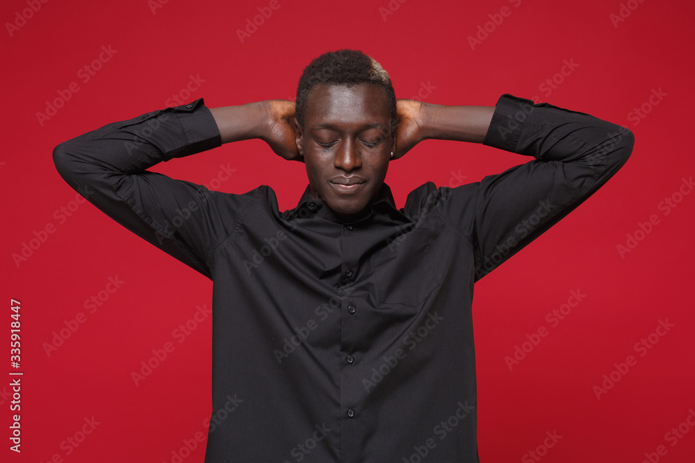 Young african american man guy in classic black shirt posing isolated on red wall background studio portrait. People emotions lifestyle concept. Mock up copy space. Sleeping with hands behind head. Young african american man guy in classic black shirt posing isolated on red wall background studio portrait. People emotions lifestyle concept. Mock up copy space. Sleeping with hands behind head.