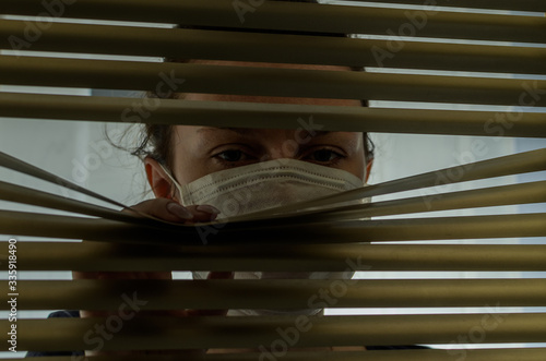 Young woman in medical mask looks out the window through the blinds