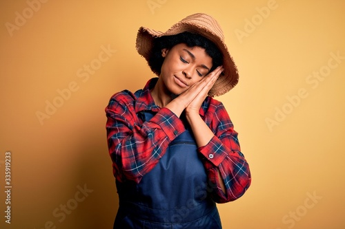 Young African American afro farmer woman with curly hair wearing apron and hat sleeping tired dreaming and posing with hands together while smiling with closed eyes.