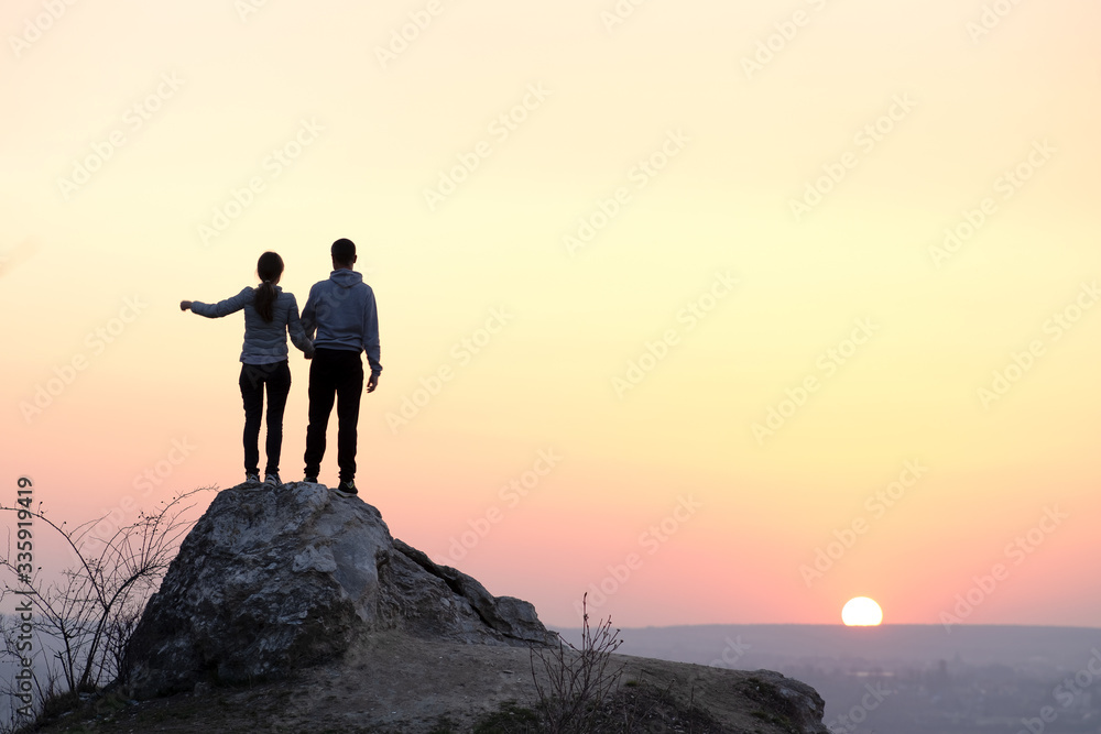 Man and woman hikers standing on big stone at sunset in mountains. Couple on high rock in evening nature. Tourism, traveling and healthy lifestyle concept.