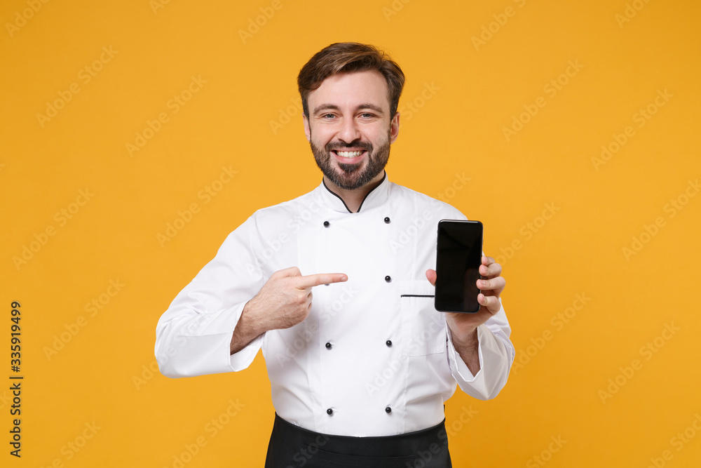 Smiling bearded male chef cook or baker man in white uniform shirt isolated on yellow background. Cooking food concept. Mock up copy space. Point index finger on mobile phone with blank empty screen. Smiling bearded male chef cook or baker man in white uniform shirt isolated on yellow background. Cooking food concept. Mock up copy space. Point index finger on mobile phone with blank empty screen.