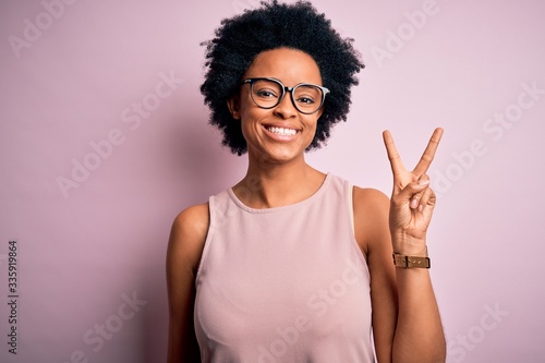 Young beautiful African American afro woman with curly hair wearing t-shirt and glasses smiling with happy face winking at the camera doing victory sign. Number two.