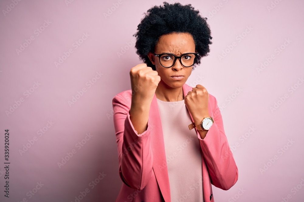 Young beautiful African American afro businesswoman with curly hair wearing pink jacket Ready to fight with fist defense gesture, angry and upset face, afraid of problem Young beautiful African American afro businesswoman with curly hair wearing pink jacket Ready to fight with fist defense gesture, angry and upset face, afraid of problem