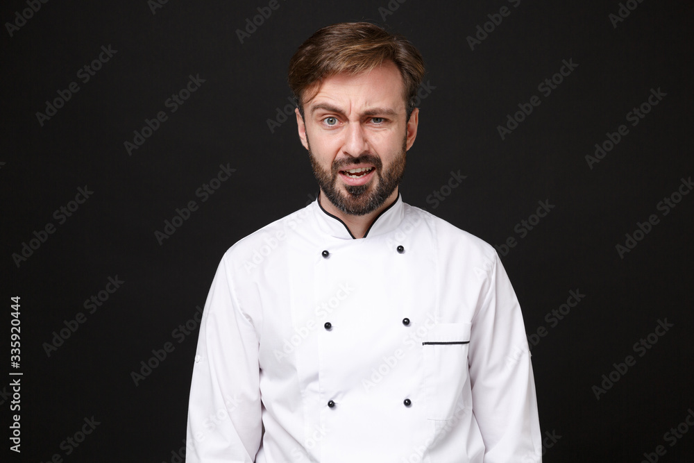 Confused perplexed young bearded male chef cook or baker man in white uniform shirt posing isolated on black wall background studio portrait. Cooking food concept. Mock up copy space. Looking camera.