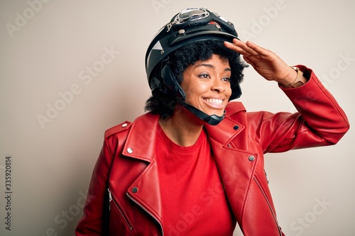 Young African American afro motorcyclist woman with curly hair wearing motorcycle helmet very happy and smiling looking far away with hand over head. Searching concept.