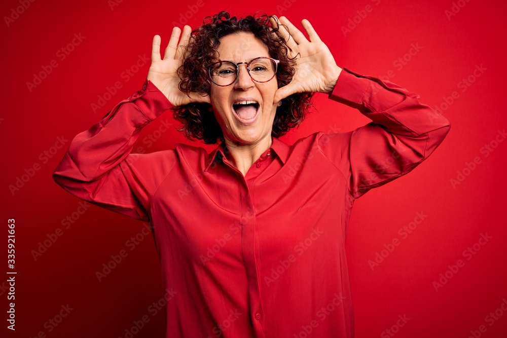Middle age beautiful curly hair woman wearing casual shirt and glasses over red background Smiling cheerful playing peek a boo with hands showing face. Surprised and exited Middle age beautiful curly hair woman wearing casual shirt and glasses over red background Smiling cheerful playing peek a boo with hands showing face. Surprised and exited