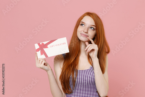 Pensive young redhead woman girl in plaid dress posing isolated on pastel pink background studio portrait. People lifestyle concept. Mock up copy space. Hold gift certificate put hand prop up on chin.