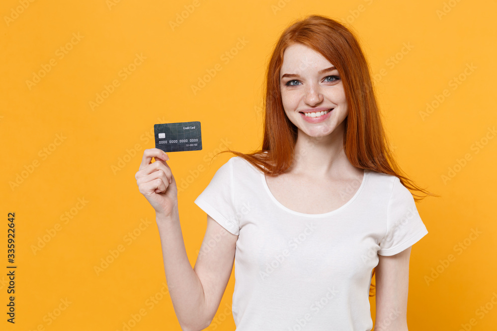 Smiling young redhead woman girl in white blank empty t-shirt posing isolated on yellow wall background studio portrait. People emotions lifestyle concept. Mock up copy space. Hold credit bank card. Smiling young redhead woman girl in white blank empty t-shirt posing isolated on yellow wall background studio portrait. People emotions lifestyle concept. Mock up copy space. Hold credit bank card.