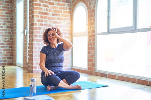 Middle age beautiful sportswoman wearing sportswear sitting on mat practicing yoga at home doing ok gesture with hand smiling, eye looking through fingers with happy face.