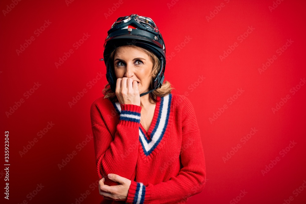 Obraz premium Middle age beautiful blonde motorcyclist woman wearing moto helmet over red background looking stressed and nervous with hands on mouth biting nails. Anxiety problem.