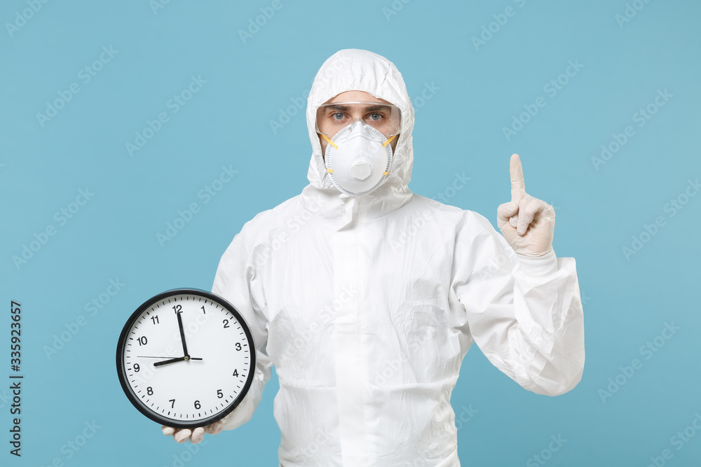 Man in white protective suit respirator mask hold in hand clock isolated on blue background studio. Epidemic pandemic new rapidly spreading coronavirus 2019-ncov from China medicine flu virus concept. Man in white protective suit respirator mask hold in hand clock isolated on blue background studio. Epidemic pandemic new rapidly spreading coronavirus 2019-ncov from China medicine flu virus concept.