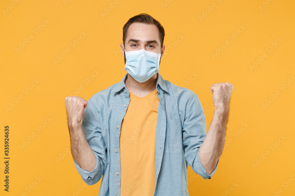 Happy young man in sterile face mask posing isolated on yellow wall background studio portrait. Epidemic pandemic spreading coronavirus 2019-ncov sars covid-19 flu virus concept. Doing winner gesture. Happy young man in sterile face mask posing isolated on yellow wall background studio portrait. Epidemic pandemic spreading coronavirus 2019-ncov sars covid-19 flu virus concept. Doing winner gesture.