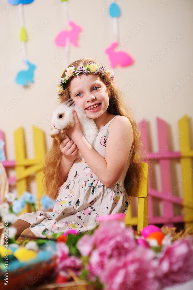 child-a little girl in a dress and a flower wreath on her head smiling and holding an Easter Bunny on the background of flowers and decor child-a little girl in a dress and a flower wreath on her head smiling and holding an Easter Bunny on the background of flowers and decor