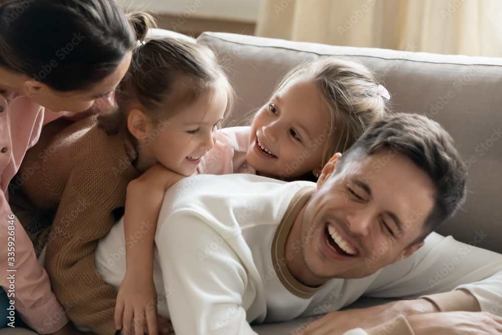 Head shot joyful bonding family of four embracing having fun together on cozy couch in living room. Laughing father resting on sofa with two funny cute kids daughters and smiling wife on back. Head shot joyful bonding family of four embracing having fun together on cozy couch in living room. Laughing father resting on sofa with two funny cute kids daughters and smiling wife on back.
