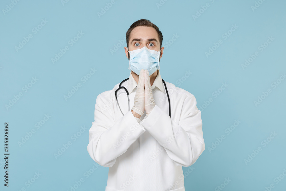 Male doctor man in white medical gown sterile face mask gloves isolated on blue background. Epidemic pandemic coronavirus 2019-ncov sars covid-19 flu virus concept. Holding hands folded in prayer. Male doctor man in white medical gown sterile face mask gloves isolated on blue background. Epidemic pandemic coronavirus 2019-ncov sars covid-19 flu virus concept. Holding hands folded in prayer.