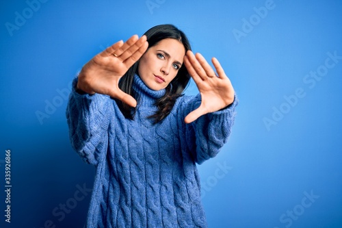 Young brunette woman with blue eyes wearing casual turtleneck sweater doing frame using hands palms and fingers, camera perspective © Krakenimages.com