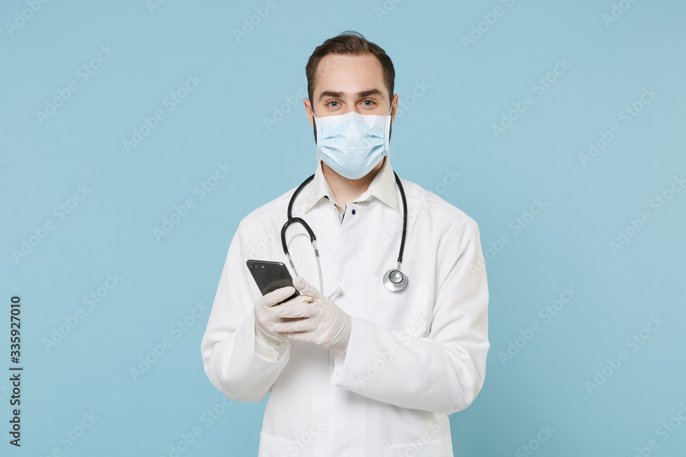 Male doctor man in medical gown sterile face mask gloves isolated on blue background. Epidemic pandemic coronavirus 2019-ncov sars covid-19 flu virus concept. Using mobile phone for on-line treatment. Male doctor man in medical gown sterile face mask gloves isolated on blue background. Epidemic pandemic coronavirus 2019-ncov sars covid-19 flu virus concept. Using mobile phone for on-line treatment.