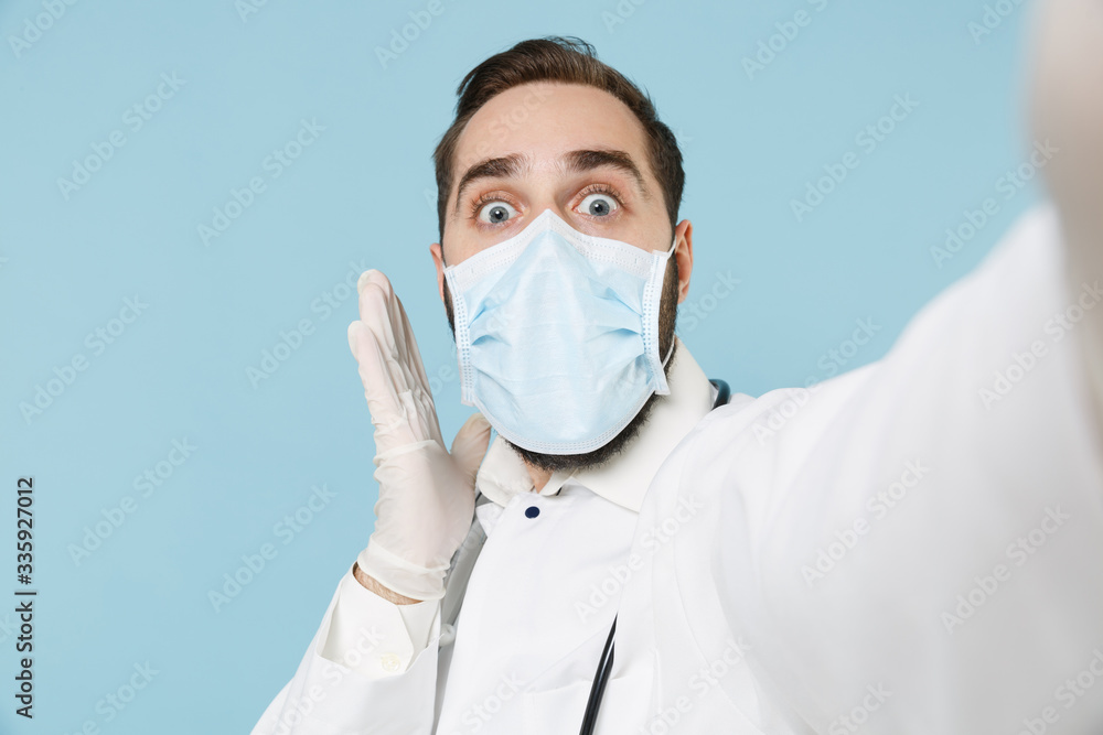 Close up of shocked male doctor man in medical gown face mask gloves isolated on blue background. Epidemic pandemic coronavirus 2019-ncov sars covid-19 flu virus concept. Doing selfie on mobile phone. Close up of shocked male doctor man in medical gown face mask gloves isolated on blue background. Epidemic pandemic coronavirus 2019-ncov sars covid-19 flu virus concept. Doing selfie on mobile phone.