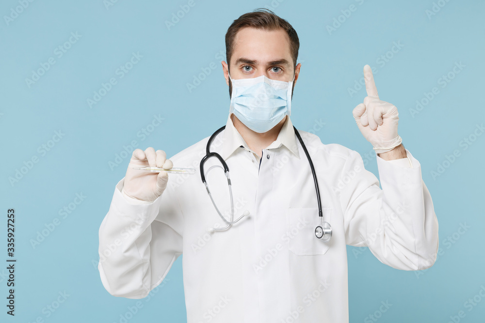Male doctor man in medical gown sterile face mask gloves isolated on blue background. Epidemic pandemic coronavirus 2019-ncov sars covid-19 flu virus concept. Hold thermometer, point index finger up.