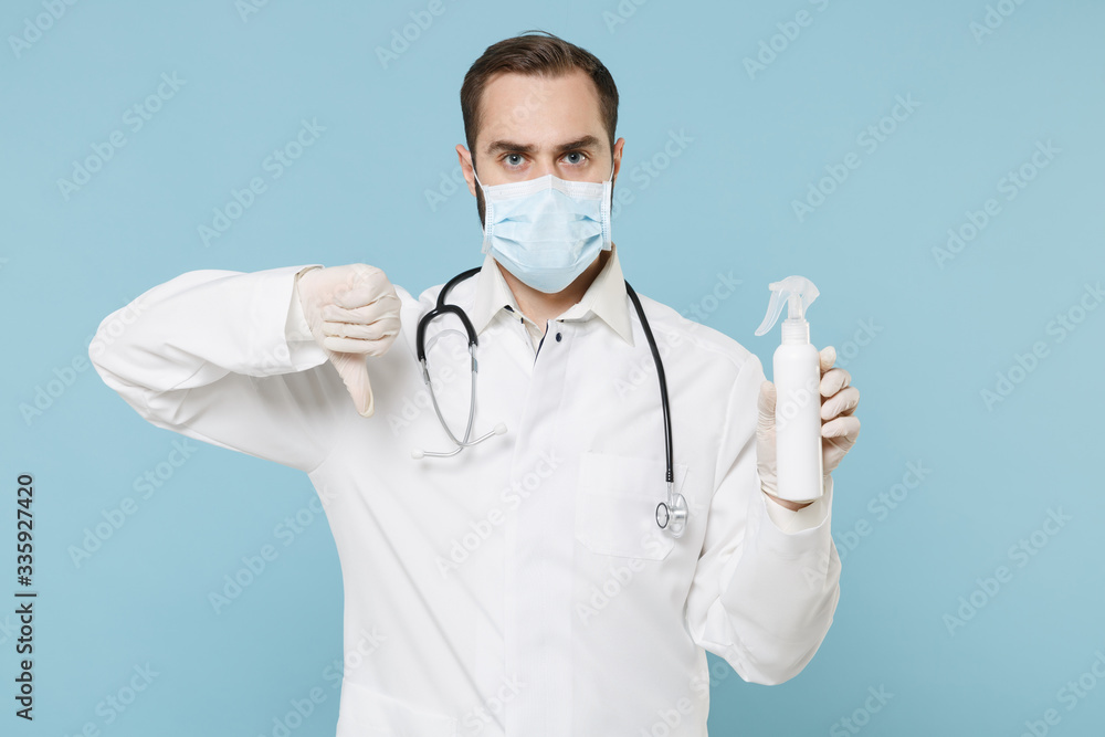 Doctor man in gown face mask gloves isolated on blue background. Epidemic pandemic coronavirus 2019-ncov sars covid-19 flu virus. Hold bottle alcohol liquid antibacterial sanitizer showing thumb down.