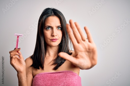 Young brunette woman with blue eyes wearing bath towel using razor over white background with open hand doing stop sign with serious and confident expression, defense gesture photo