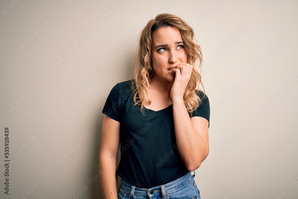 Young beautiful blonde woman wearing casual t-shirt standing over isolated white background looking stressed and nervous with hands on mouth biting nails. Anxiety problem. Young beautiful blonde woman wearing casual t-shirt standing over isolated white background looking stressed and nervous with hands on mouth biting nails. Anxiety problem.
