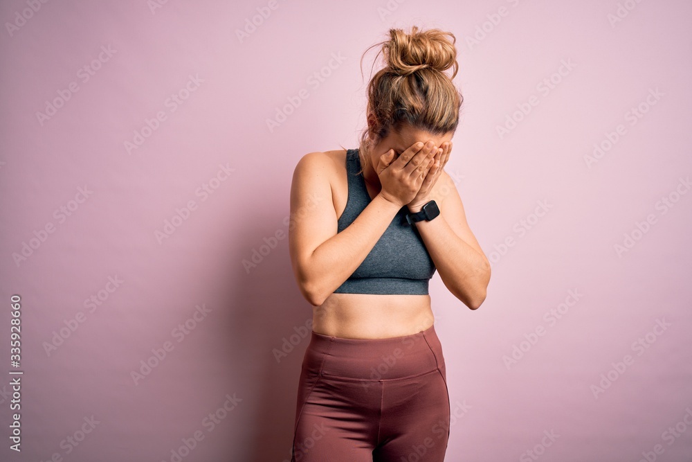 Young beautiful blonde sportswoman doing sport wearing sportswear over pink background with sad expression covering face with hands while crying. Depression concept.
