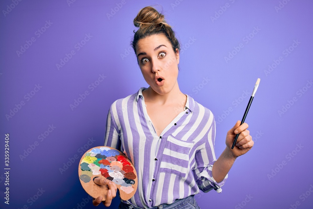 Fototapeta premium Young beautiful blonde artist woman painting using brush and palette over purple background scared in shock with a surprise face, afraid and excited with fear expression