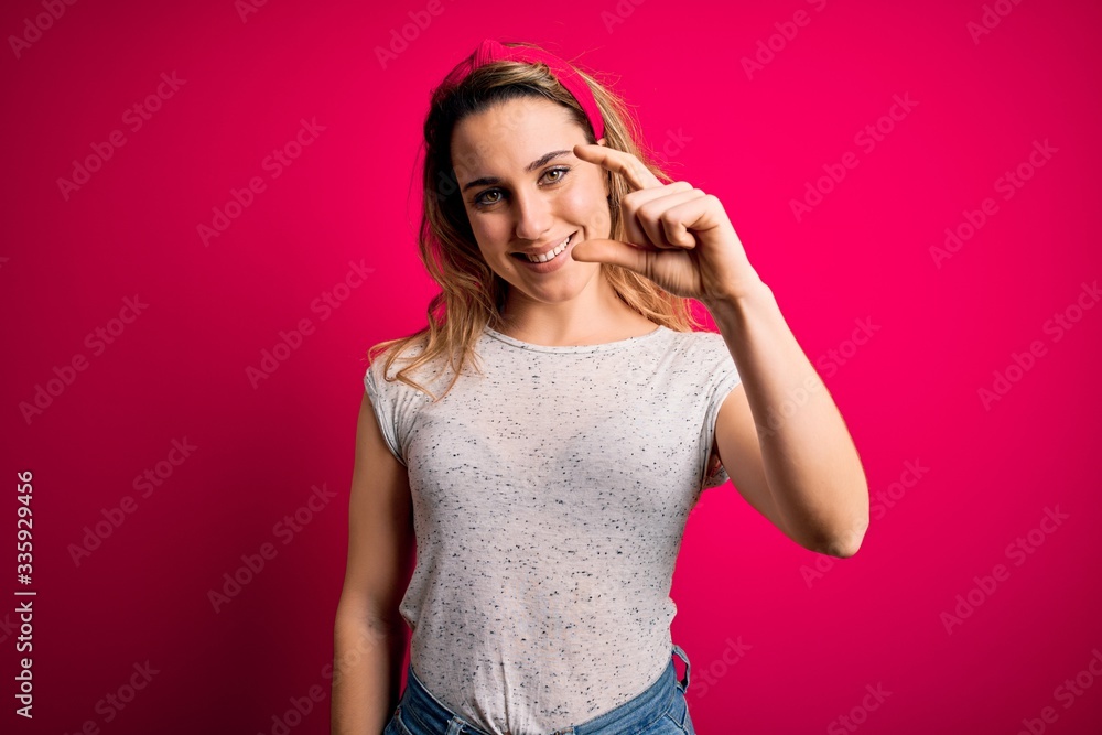 Young beautiful blonde woman wearing casual t-shirt standing over isolated pink background smiling and confident gesturing with hand doing small size sign with fingers looking and the camera. Measure Young beautiful blonde woman wearing casual t-shirt standing over isolated pink background smiling and confident gesturing with hand doing small size sign with fingers looking and the camera. Measure
