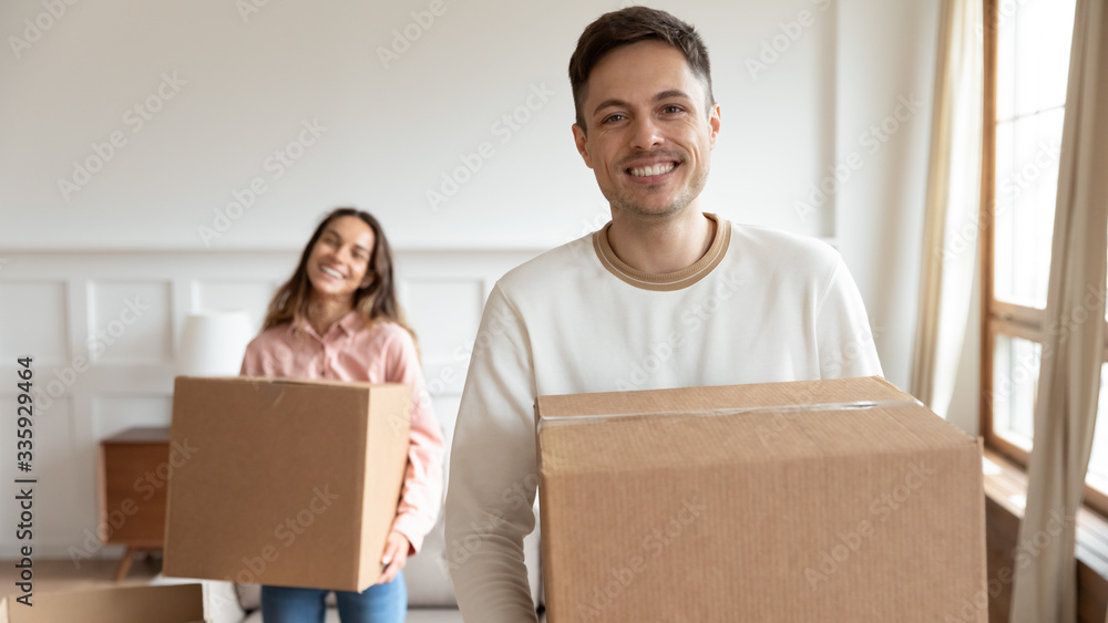 Focus on happy young husband holding box with belongings with blurred mixed race wife holding carton parcel on background. Smiling family couple moving in new apartment, real estate concept. Focus on happy young husband holding box with belongings with blurred mixed race wife holding carton parcel on background. Smiling family couple moving in new apartment, real estate concept.