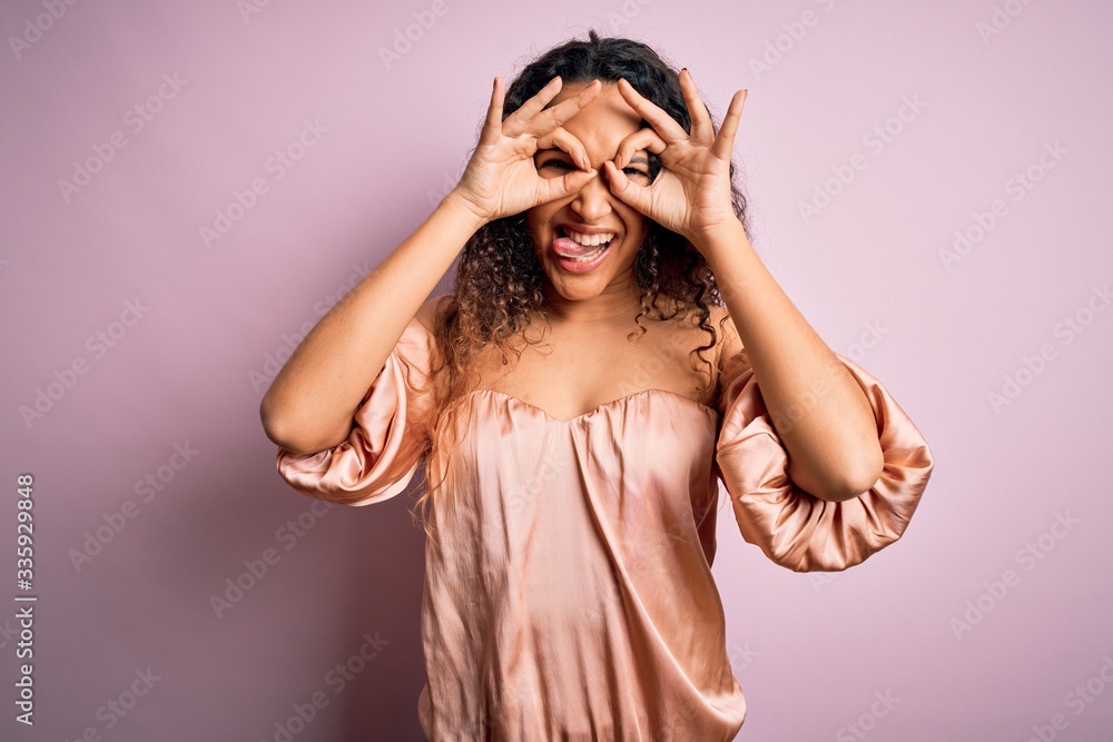 Young beautiful woman with curly hair wearing casual t-shirt standing over pink background doing ok gesture like binoculars sticking tongue out, eyes looking through fingers. Crazy expression. Young beautiful woman with curly hair wearing casual t-shirt standing over pink background doing ok gesture like binoculars sticking tongue out, eyes looking through fingers. Crazy expression.