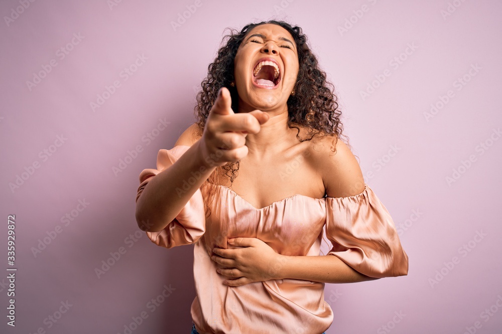 Young beautiful woman with curly hair wearing casual t-shirt standing over pink background laughing at you, pointing finger to the camera with hand over body, shame expression Young beautiful woman with curly hair wearing casual t-shirt standing over pink background laughing at you, pointing finger to the camera with hand over body, shame expression