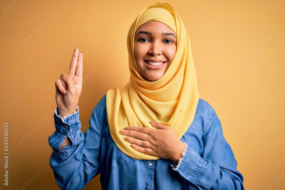 Young beautiful woman with curly hair wearing arab traditional hijab over yellow background smiling swearing with hand on chest and fingers up, making a loyalty promise oath Young beautiful woman with curly hair wearing arab traditional hijab over yellow background smiling swearing with hand on chest and fingers up, making a loyalty promise oath