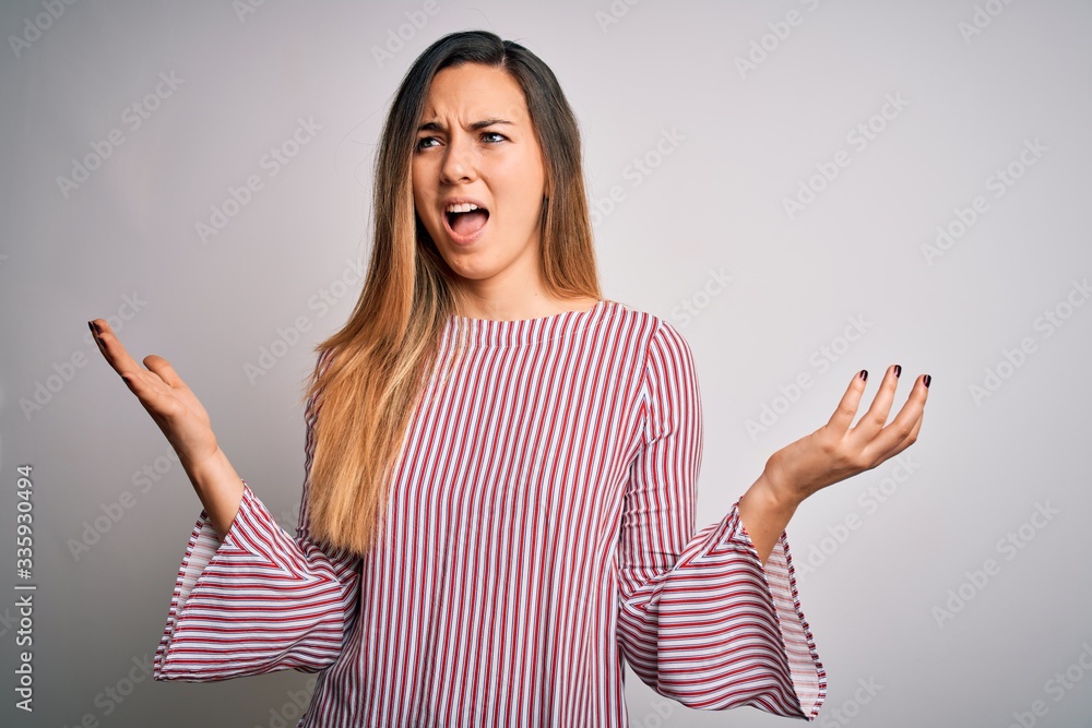 Young beautiful blonde woman with blue eyes wearing stiped t-shirt over white background crazy and mad shouting and yelling with aggressive expression and arms raised. Frustration concept. Young beautiful blonde woman with blue eyes wearing stiped t-shirt over white background crazy and mad shouting and yelling with aggressive expression and arms raised. Frustration concept.